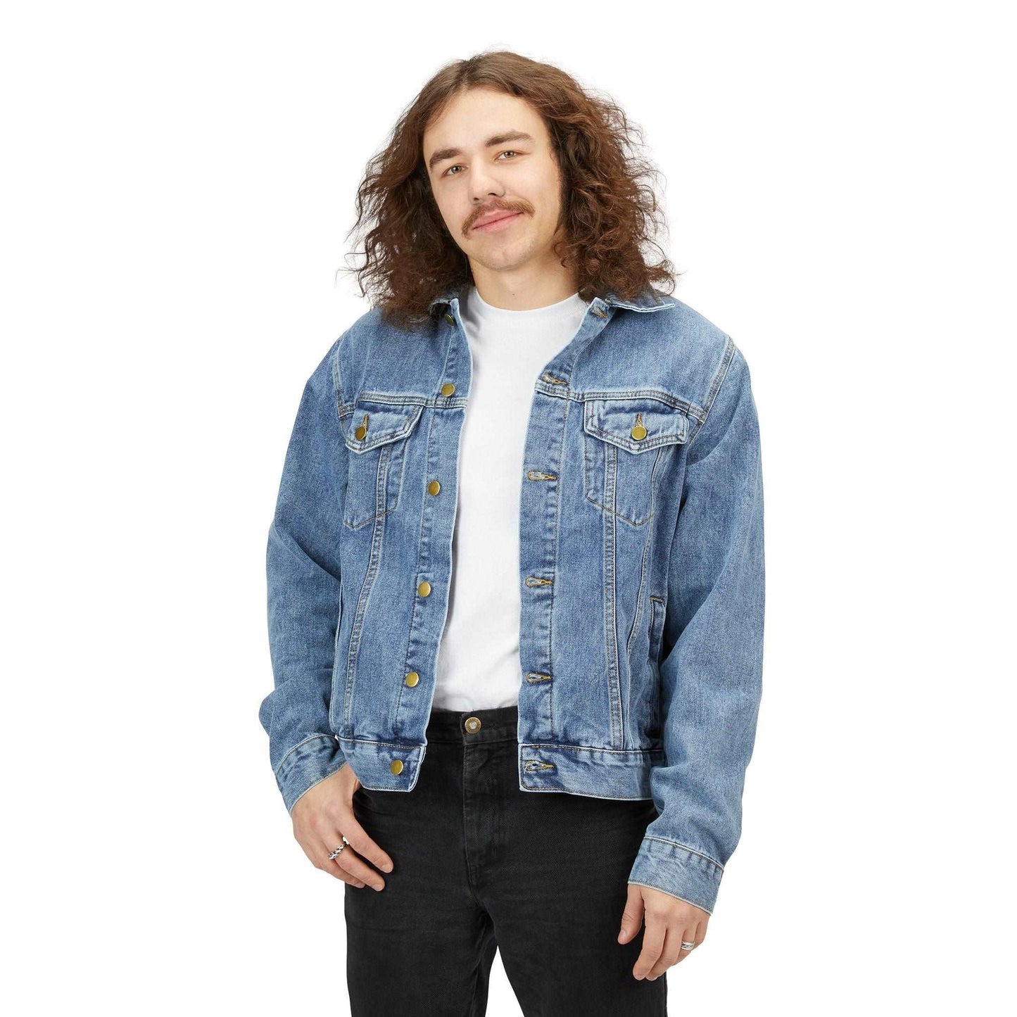 A person with long curly hair is wearing a denim jacket over a white shirt and dark pants, standing against a plain white background. They have a relaxed posture and are smiling gently.