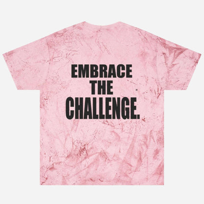 A pink tie-dye t-shirt with a textured pattern reminiscent of a medieval shirt, featuring a motivational message in bold black letters that reads "Embrace the Challenge" on the back.